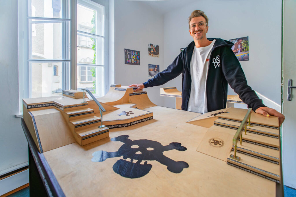 Fabi, ein junger Mann mit weißen T-Shirt und schwarzen Zip Hoodie steht vor dem G6 Plaza Fingerboard Park von Blackriver Ramps.